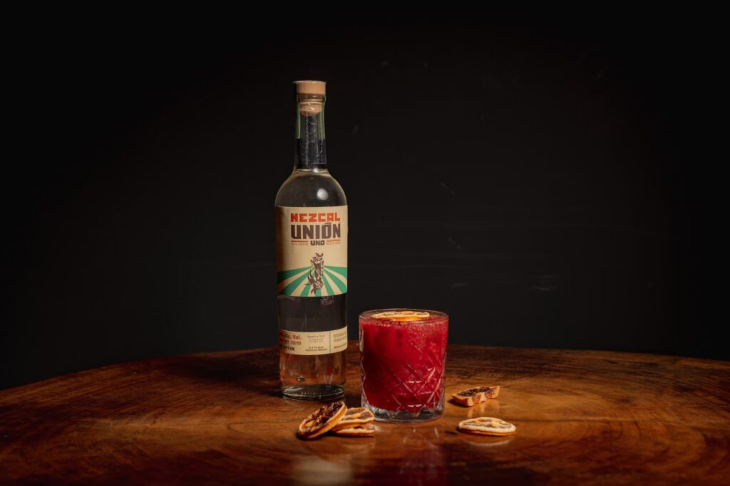A crafted mezcal cocktail garnished with dried citrus slices, sitting next to a bottle of Mezcal Union. 