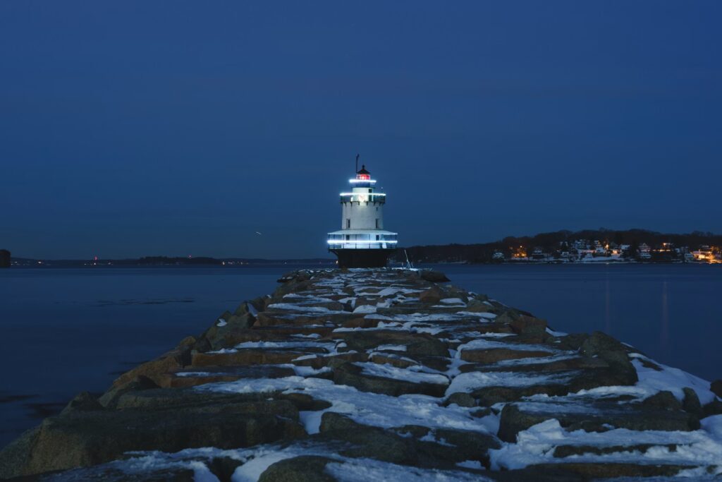 A lighthouse at dusk situated at the end of a snow-covered rocky breakwater.