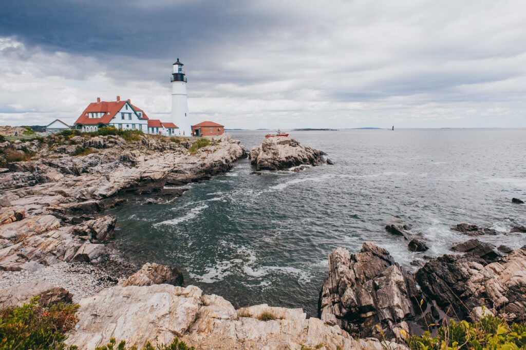 A classic white lighthouse with red-roofed buildings perched on a rugged, wave-battered rocky point.