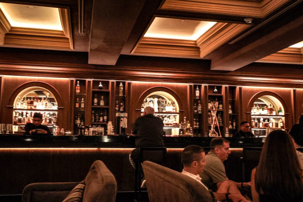 A dimly lit, sophisticated cocktail bar with patrons sitting at the counter and a well-stocked liquor display. 