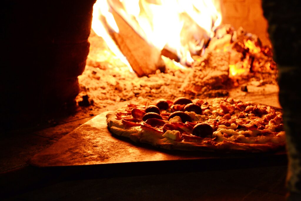 A delicious wood-fired pizza baking beside bright orange flames inside a brick oven.