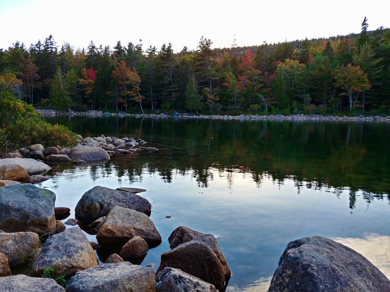 A calm lake bordered by boulders in Baxter State Park, displaying peak Maine fall foliage.