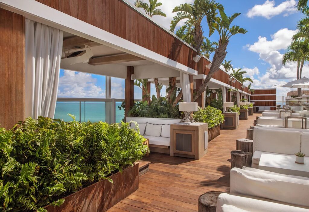A modern outdoor lounge area featuring white sofas, wooden floors, and planters, offering a view of the ocean.