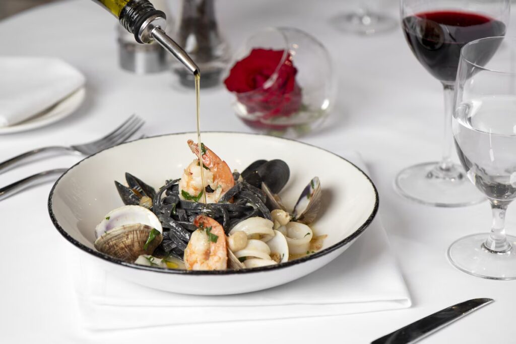 Olive oil is being poured over a gourmet dish of black squid ink pasta with shrimp and clams.