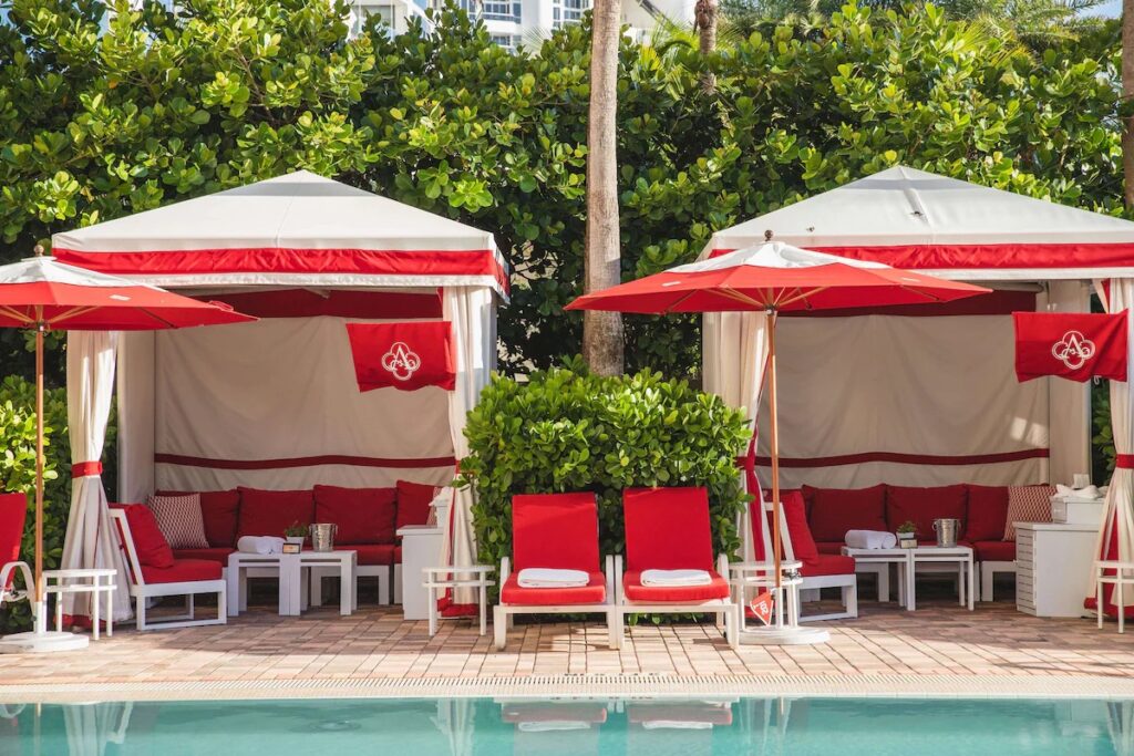 Red lounge chairs and white luxury cabanas with red trim set beside a clear swimming pool.