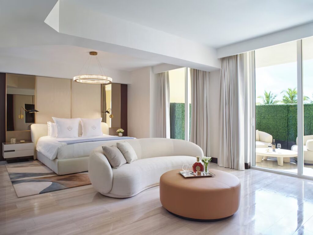 A bright, elegant suite with a large bed, curved white sofa, and floor-to-ceiling windows opening to a private patio.