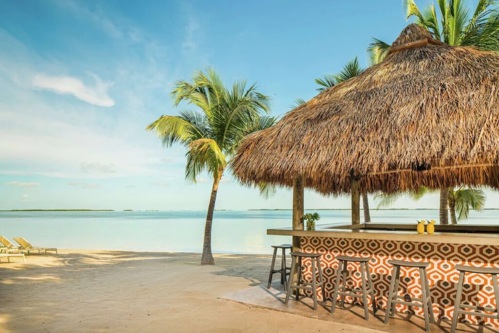 A thatched-roof beach bar with wooden barstools overlooking calm turquoise waters.