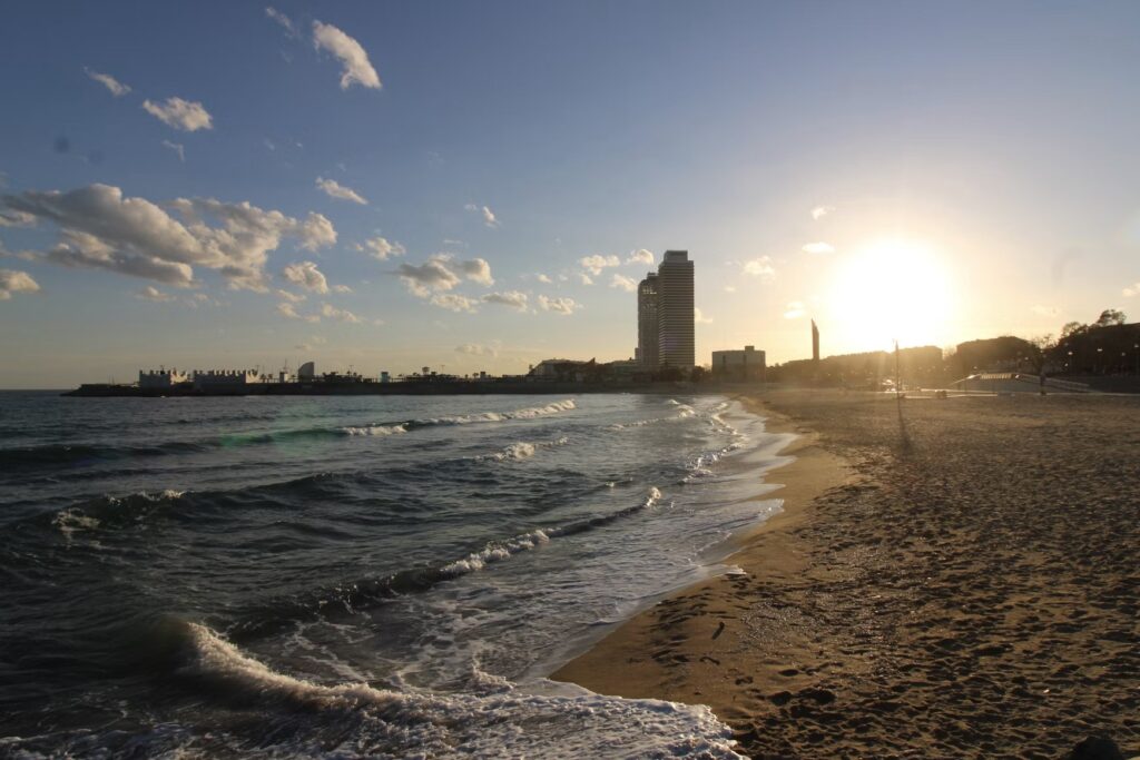 Gentle waves rolling onto a sandy beach in Barcelona as the sun sets brightly behind the city skyline.
