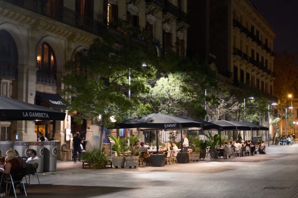 People dining at outdoor tables under black umbrellas along a wide, illuminated pedestrian street at night.