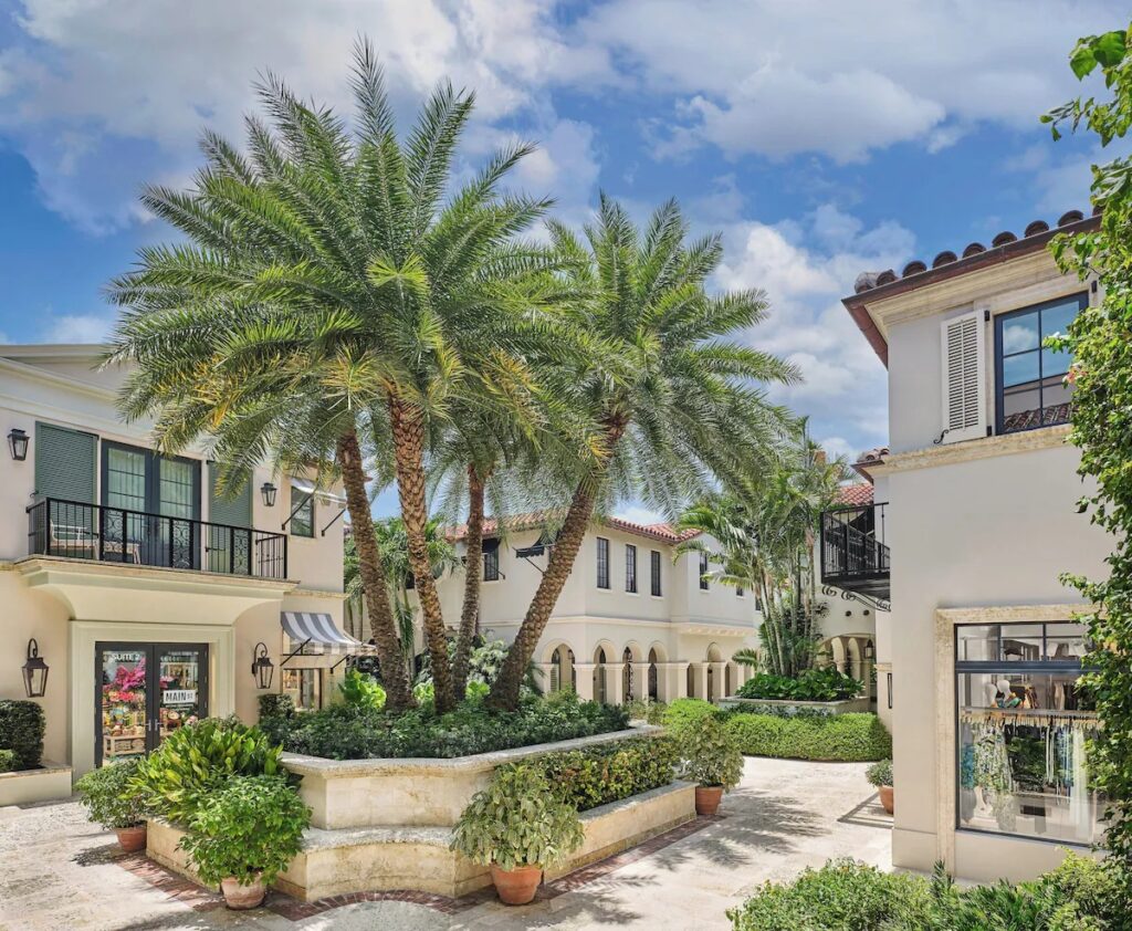 A Mediterranean-style courtyard with palm trees, stone pavers, and boutique shop entrances.