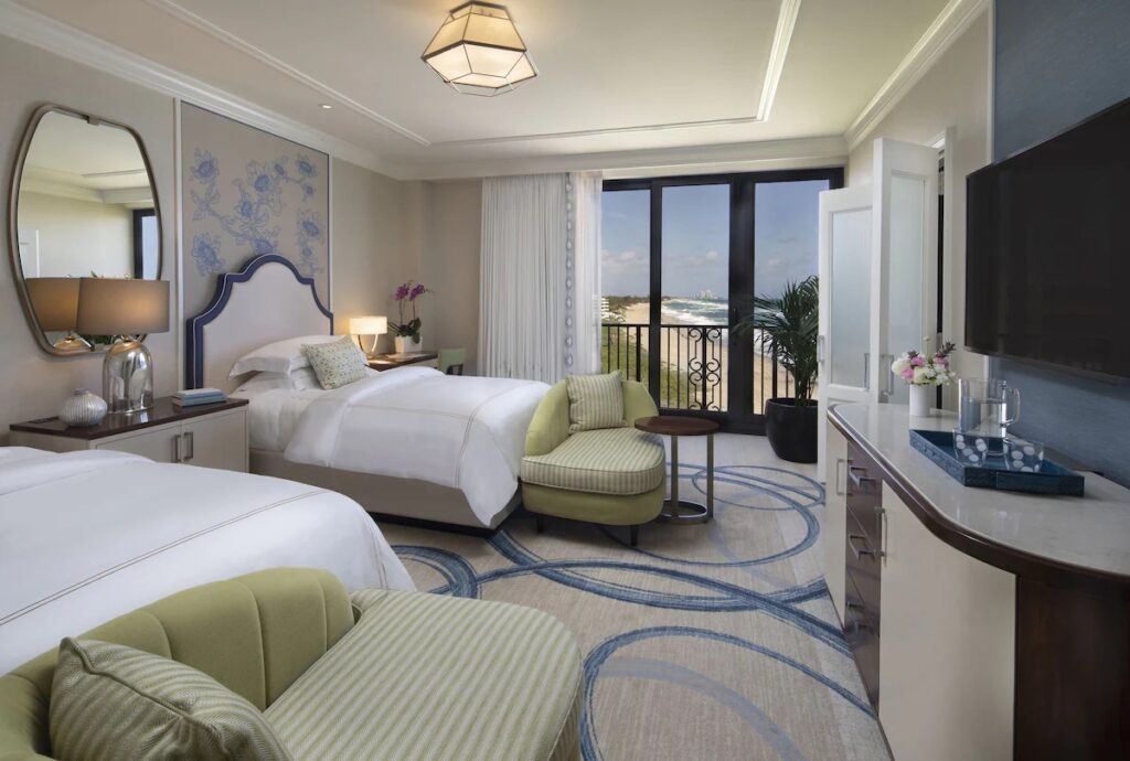 A stylish bedroom with two beds, a green armchair, and sliding glass doors to an ocean balcony.