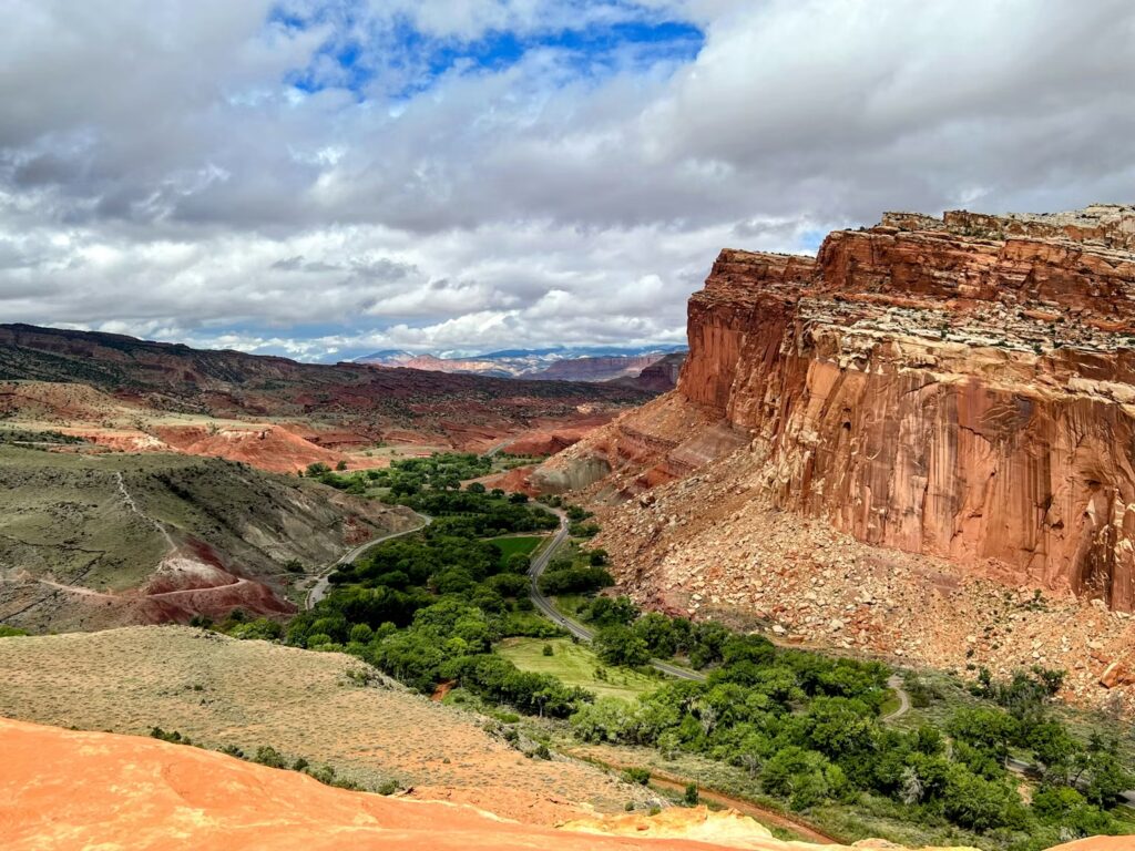 A paved road winds through a lush green valley oasis tucked between massive red rock cliffs in a desert landscape.