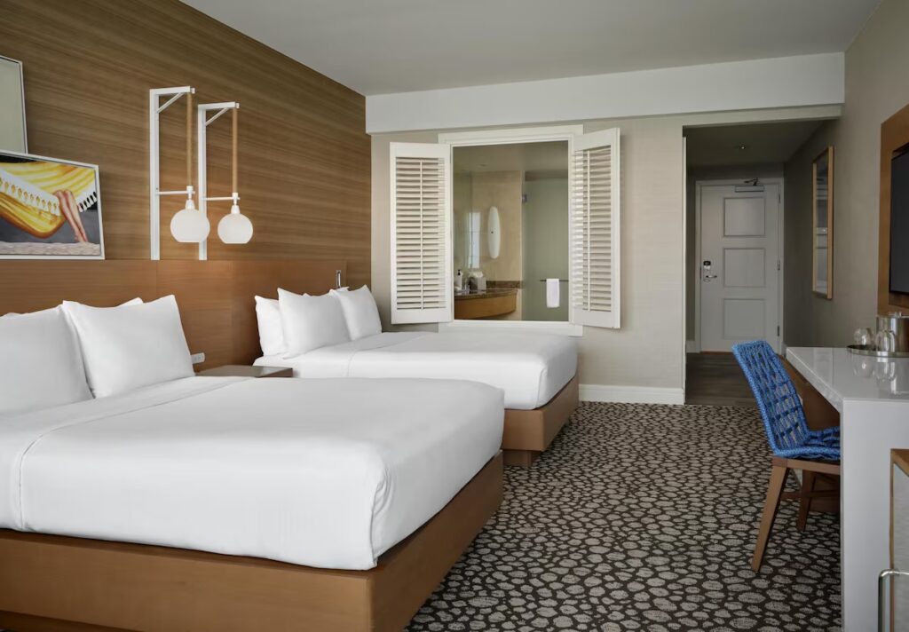 A hotel bedroom with two queen beds, wood-paneled walls, and white window shutters.