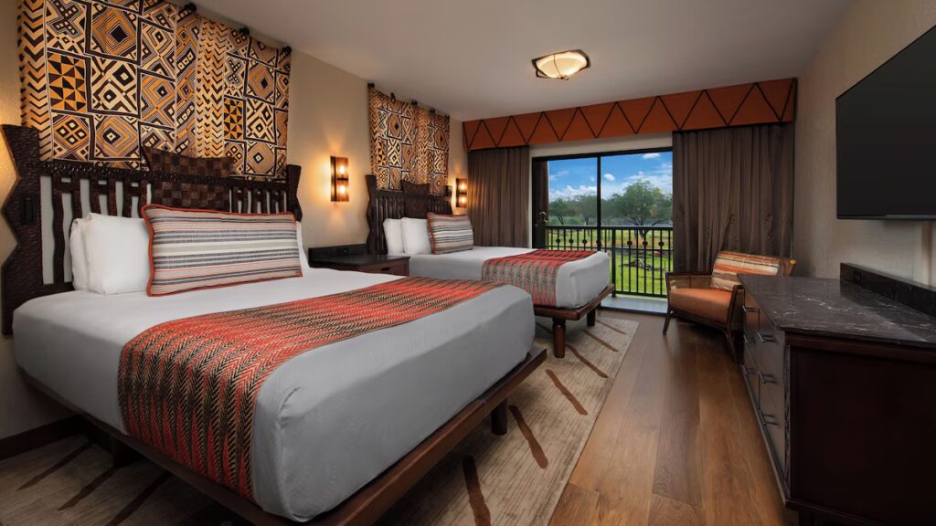 A resort bedroom with two beds, tribal pattern headboards, and a balcony view of nature.