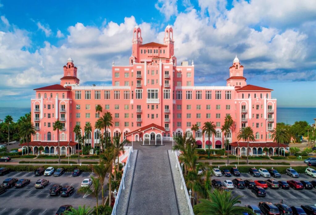 An aerial view of a large, pink multi-story hotel located right on a sandy beach next to the ocean.