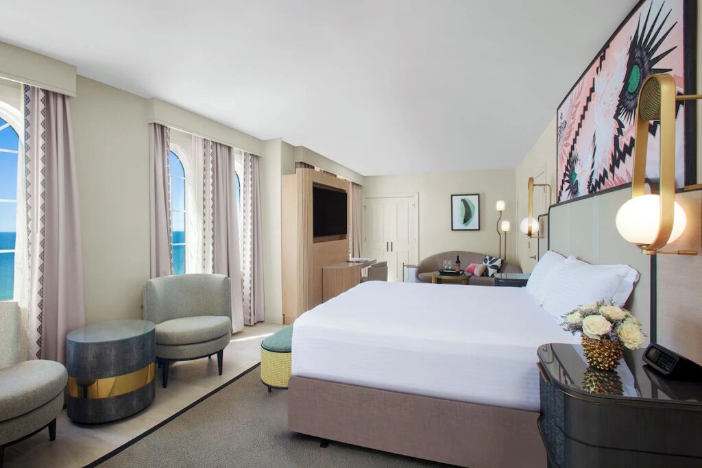 A spacious, modern hotel room featuring a king bed, a light grey sofa, and a round wooden coffee table.