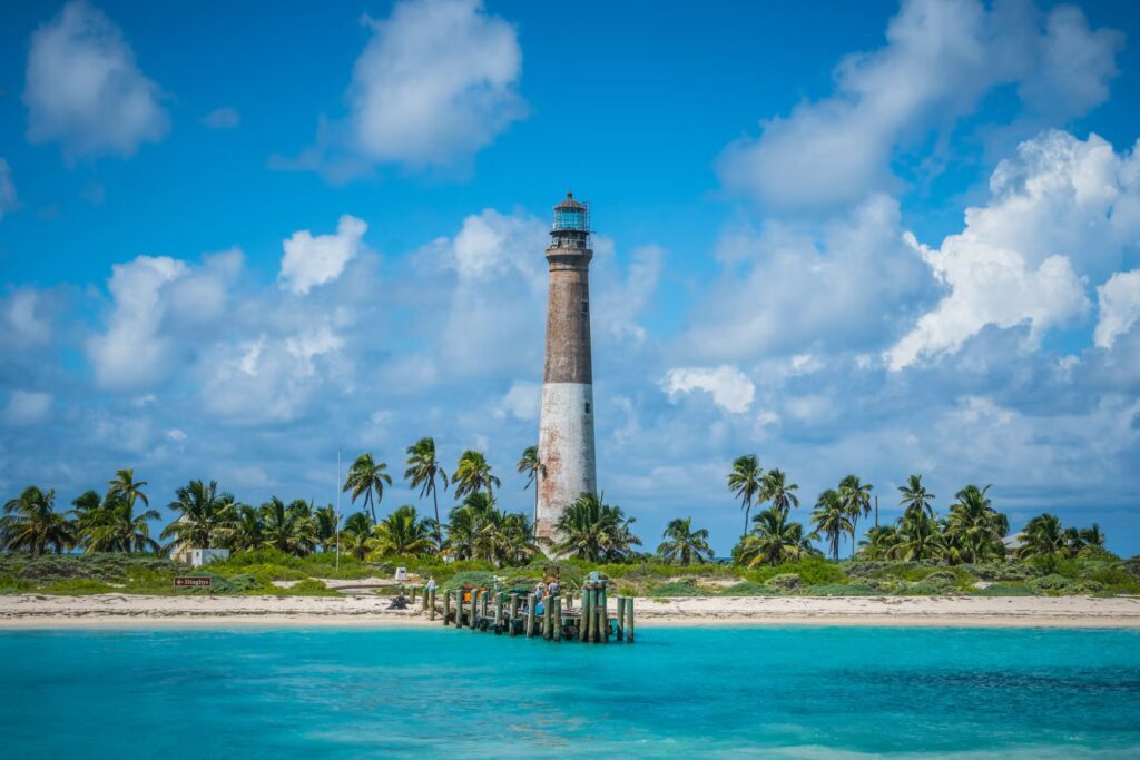 A tall, weathered brick lighthouse rises from a sandy beach lined with palm trees next to bright turquoise water.