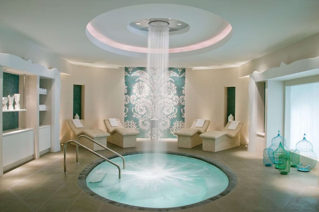 A serene spa room featuring an indoor circular plunge pool with a waterfall shower and white lounge chairs.