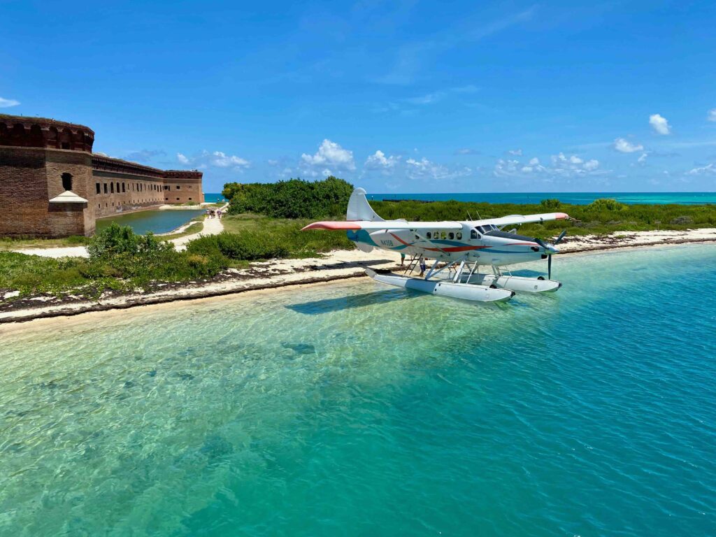 A seaplane floating in clear turquoise water near a sandy beach and the historic brick walls of Fort Jefferson.