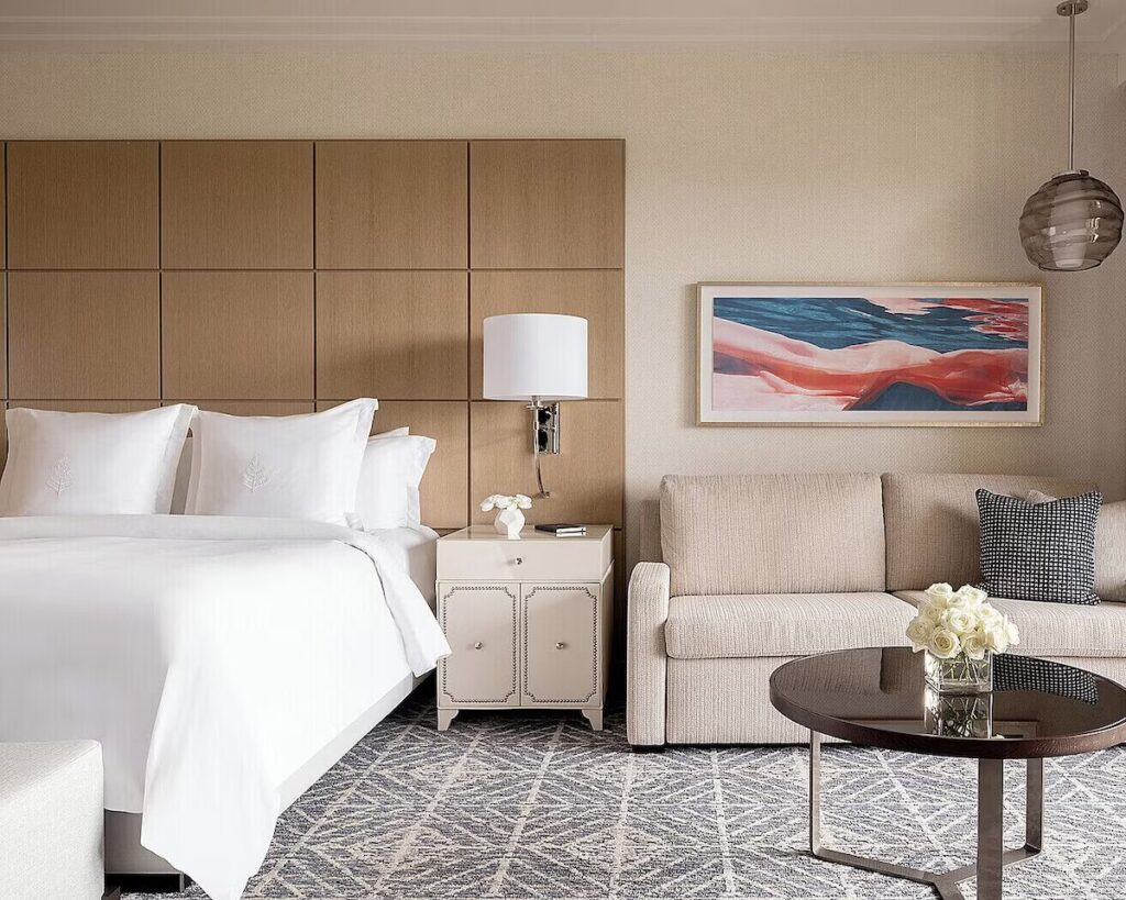 A modern hotel room with a white bed, a wooden paneled headboard, and a bright abstract painting on the wall.