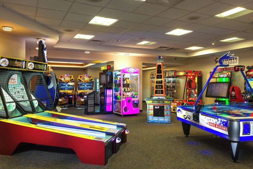 A brightly lit resort arcade room filled with basketball games, skee-ball, and claw machines.