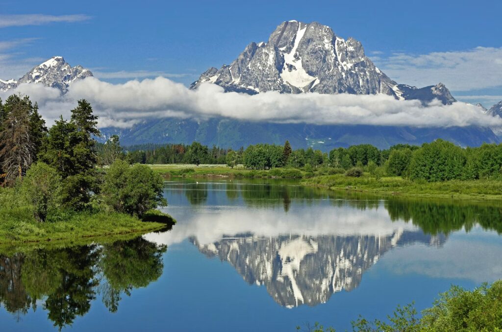 A majestic, snow-capped mountain peak reflects perfectly on the calm surface of a river surrounded by green trees.