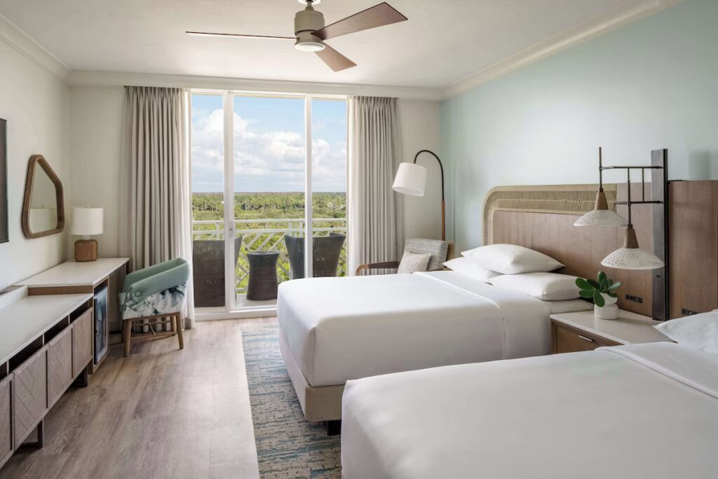 A bright guest room with two beds, light wood furniture, and a balcony overlooking a forest.