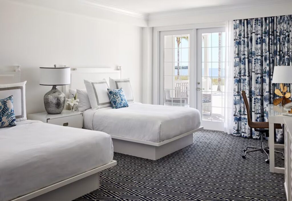 A bright bedroom with two queen beds, geometric carpet, and blue palm-patterned curtains.