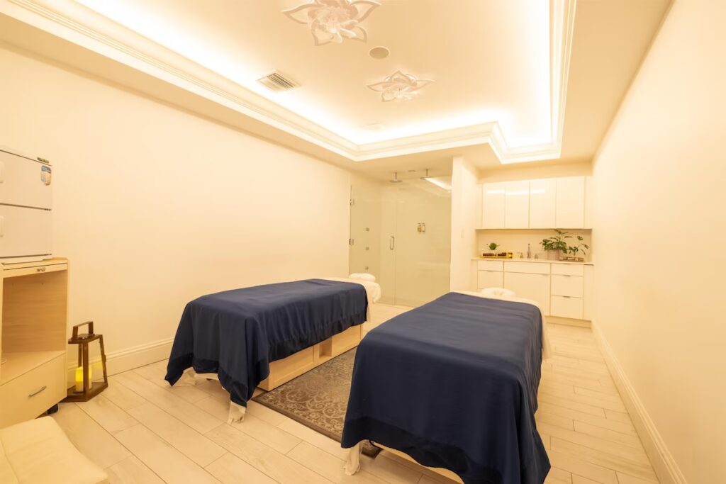 A modern spa treatment room with two massage tables covered in dark blue linens under soft lights.