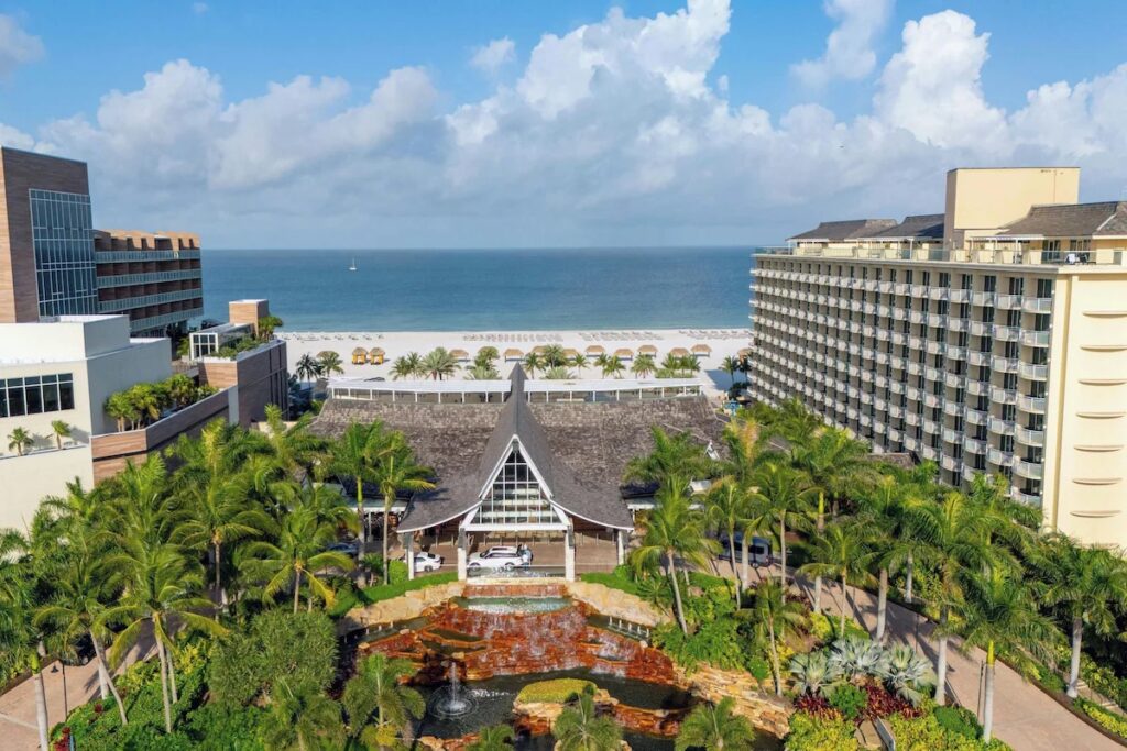 An aerial view of JW Marriott Marco Island featuring a large thatched-roof entrance, tropical pools, and the beach beyond.