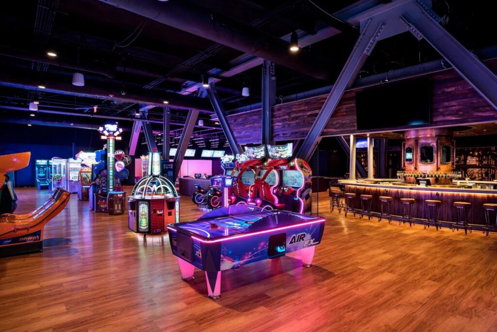 A brightly lit arcade room featuring various games, an air hockey table, and a bar area in the background.