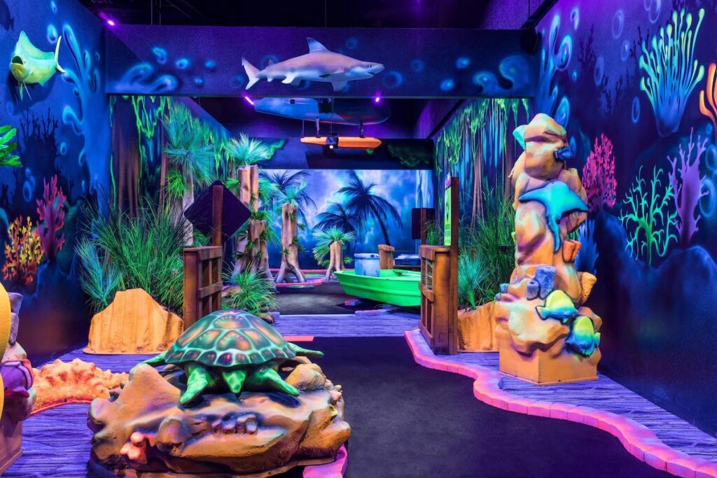 A blacklight mini-golf course decorated with a glowing sea turtle, shark, and coral reefs.