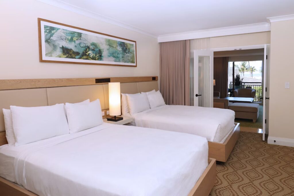 A light-filled hotel room with two queen beds opening into an adjoining seating area.