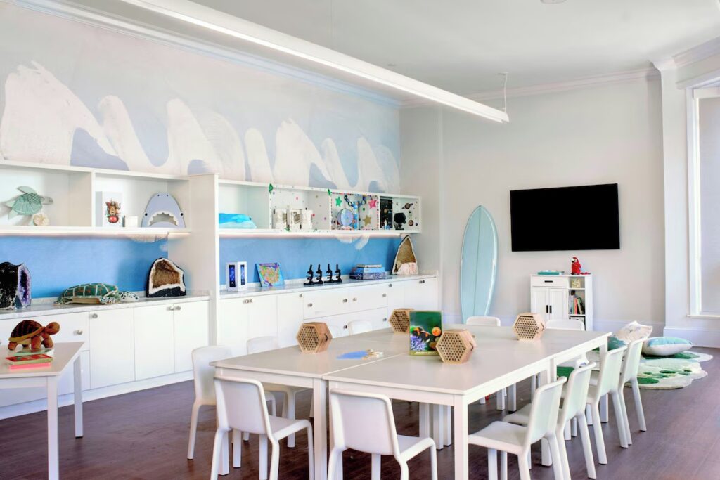 A bright children's activity room with white tables, chairs, shelving, and surfboard decor.