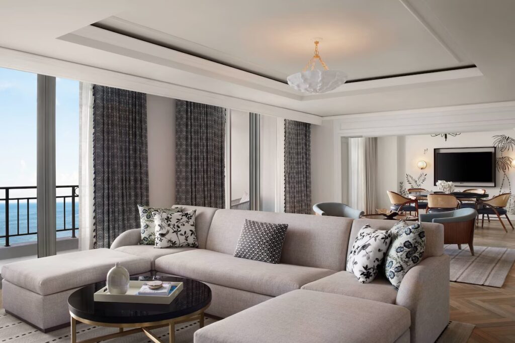 An expansive luxury suite living room with a large sofa, dining table, and ocean balcony views.