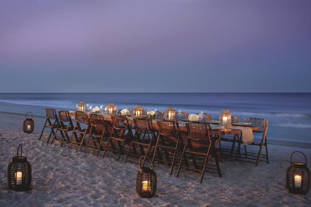A long wooden dining table set up on a sandy beach at twilight, illuminated by candles and lanterns.
