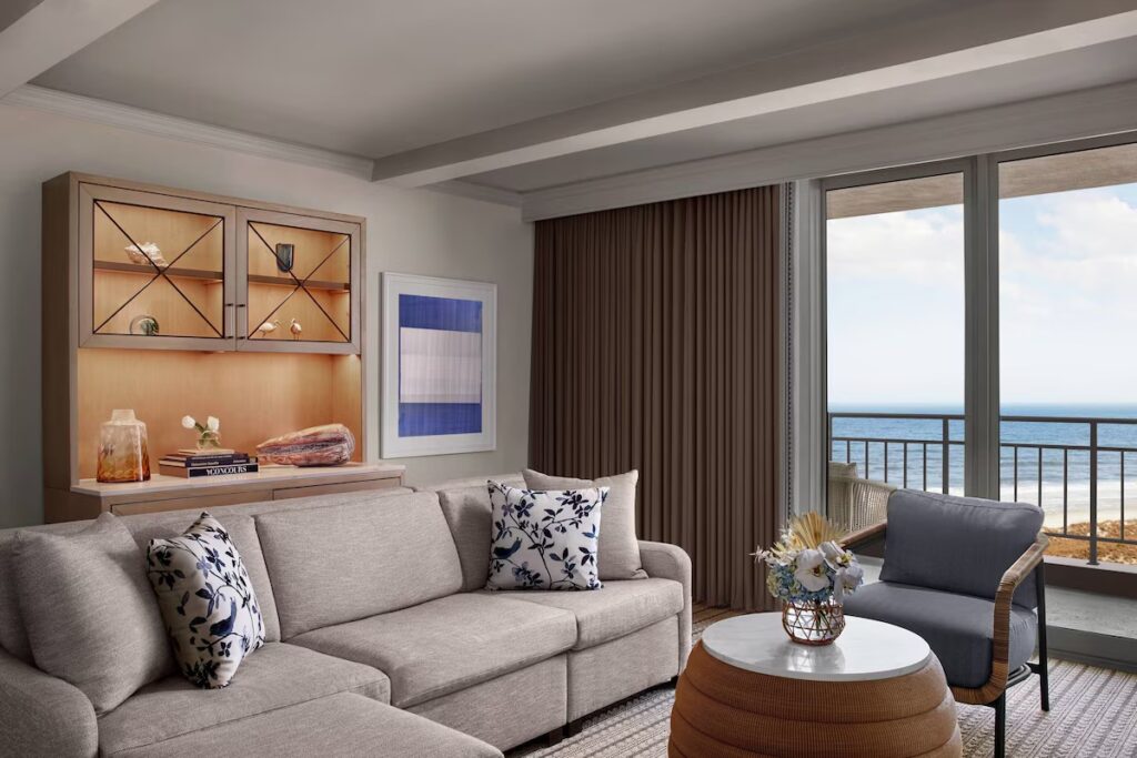 A contemporary hotel room featuring a grey sectional sofa, a round woven coffee table, and a balcony view of the beach.
