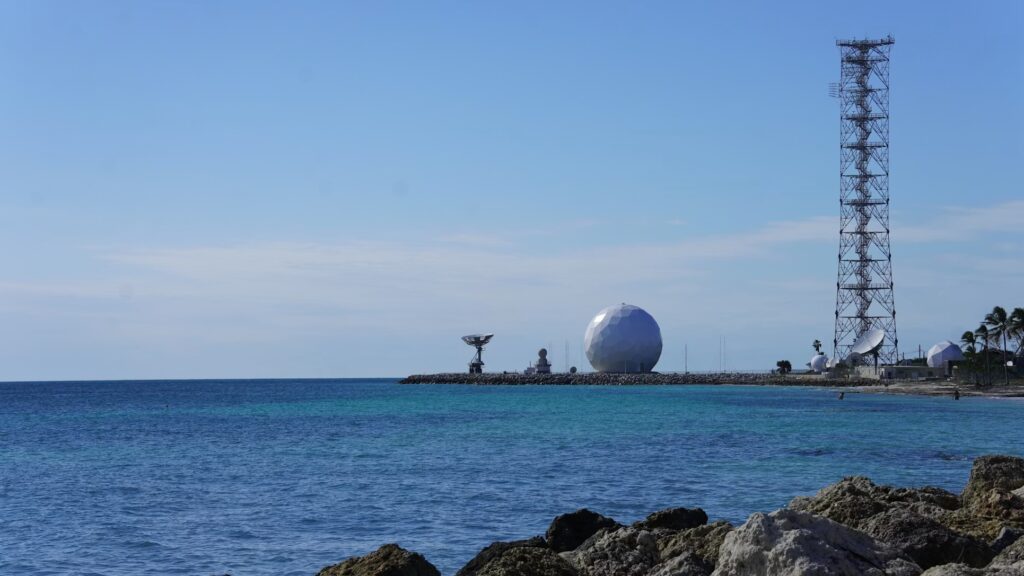 A radar dome and communication tower standing along the rocky coastline surrounded by turquoise water.