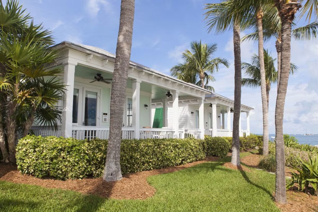 Several white cottages with porches and columns surrounded by palm trees and green bushes near the ocean.