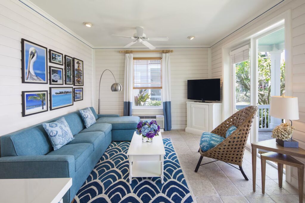 A bright, airy living room with a blue sofa, a wicker chair, and framed coastal pictures on a white wood-paneled wall.