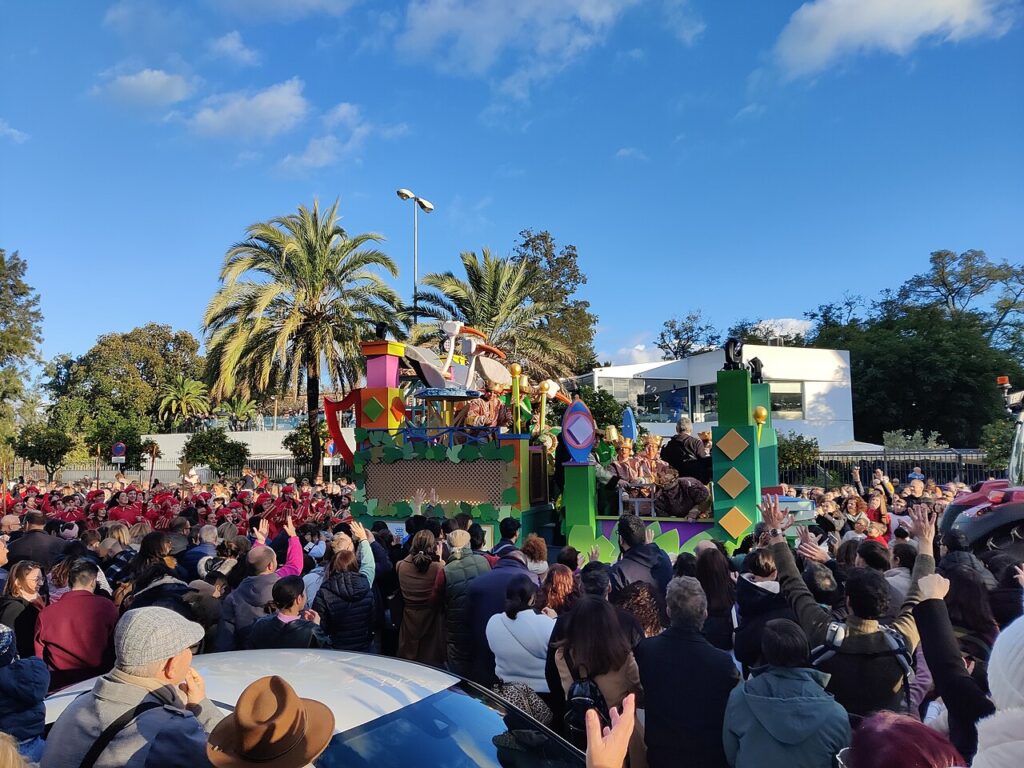 A dense crowd gathers around a colorful parade float during a sunny daytime celebration, reaching up towards the riders.