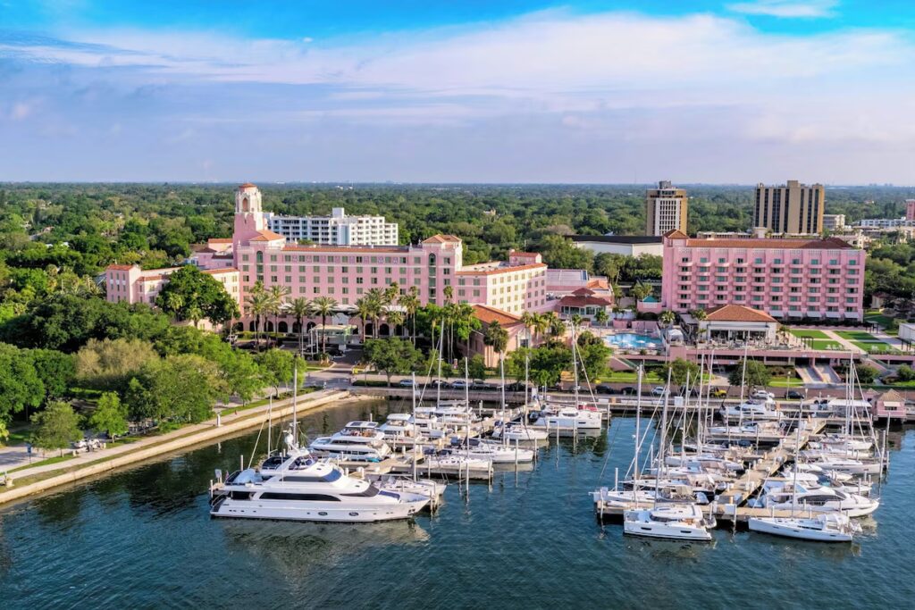 An aerial view of The Vinoy Resort showing its pink historic building, a marina filled with boats, and surrounding green trees.
