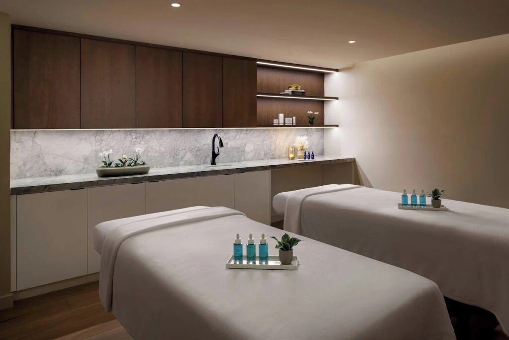 A bright, clean spa treatment room equipped with two massage tables, wooden cabinets, and subtle lighting.