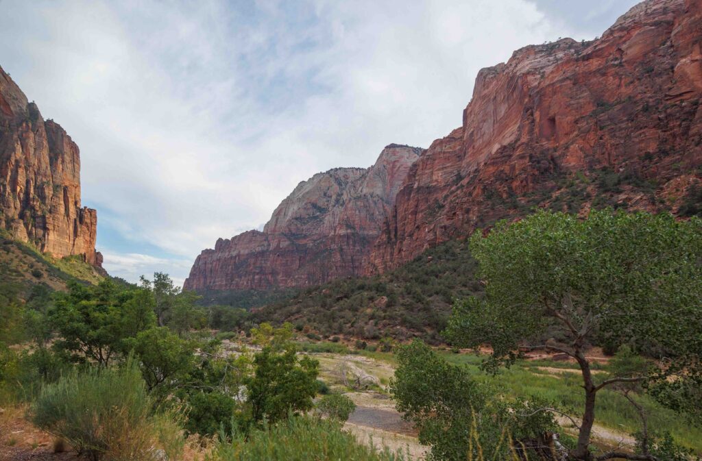 Tall, steep red sandstone cliffs rise on both sides of a valley filled with green bushes and trees.