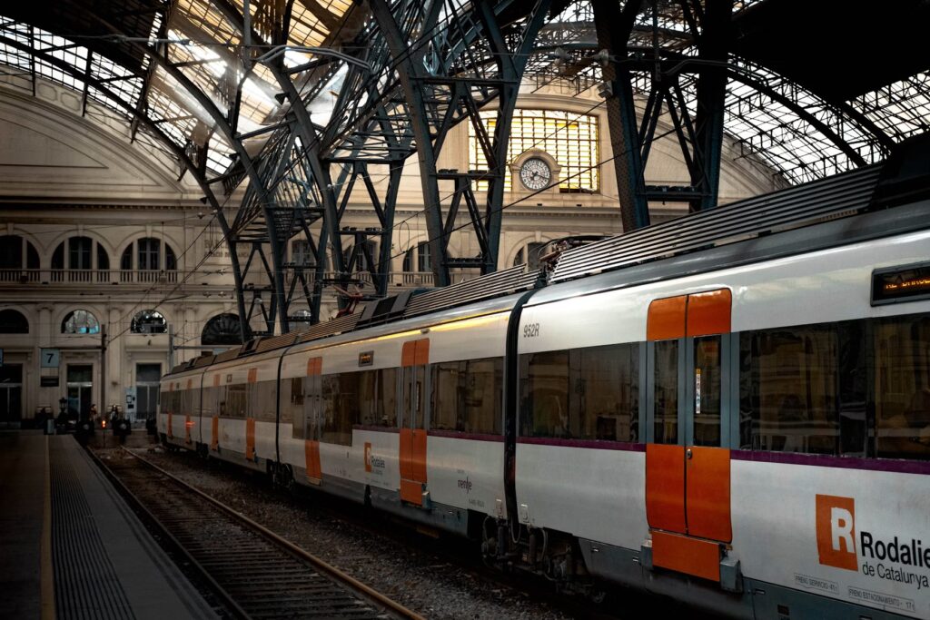 A modern Rodalies de Catalunya train waiting at a platform beneath a large, arched iron and glass station roof.