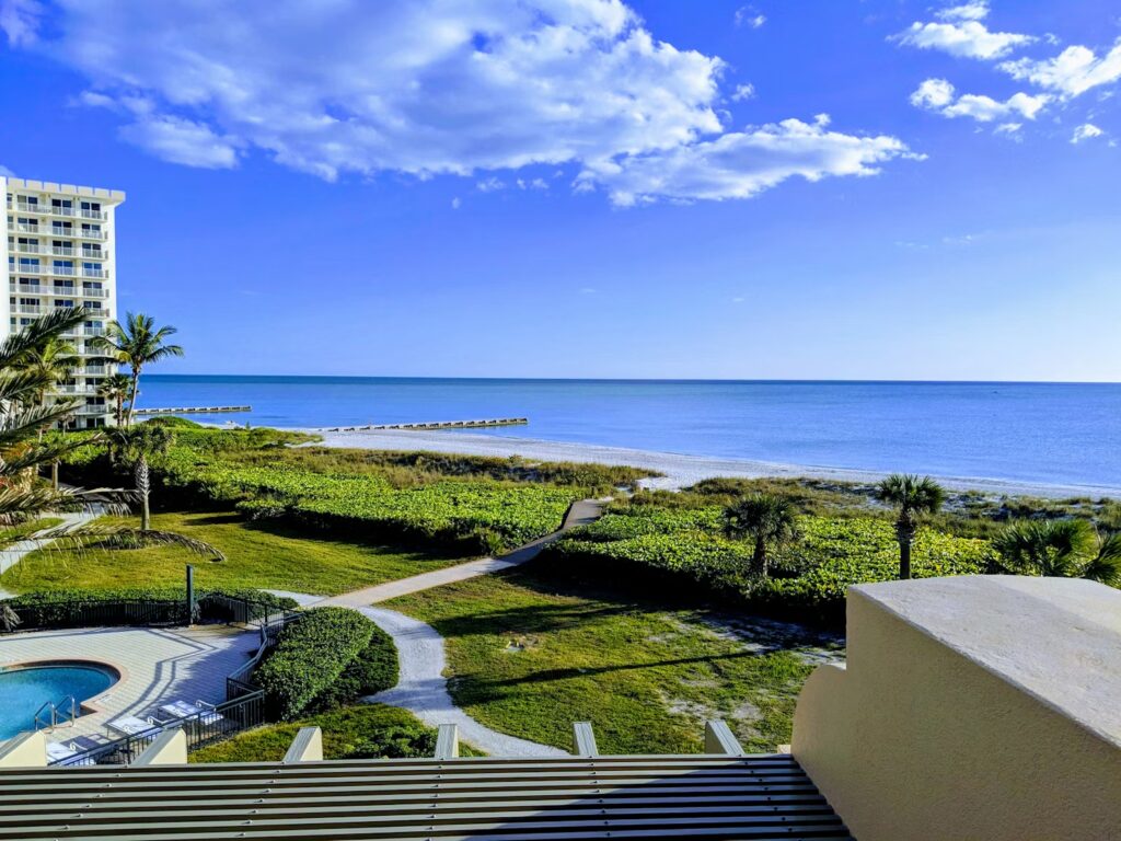 A high-angle view from a balcony showing a sandy beach, green vegetation, and calm blue ocean water.