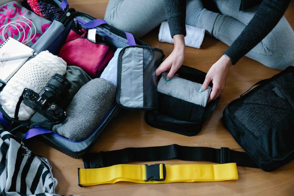 Hands organizing travel wardrobe by placing a folded gray shirt into a black mesh packing cube next to an open luggage.