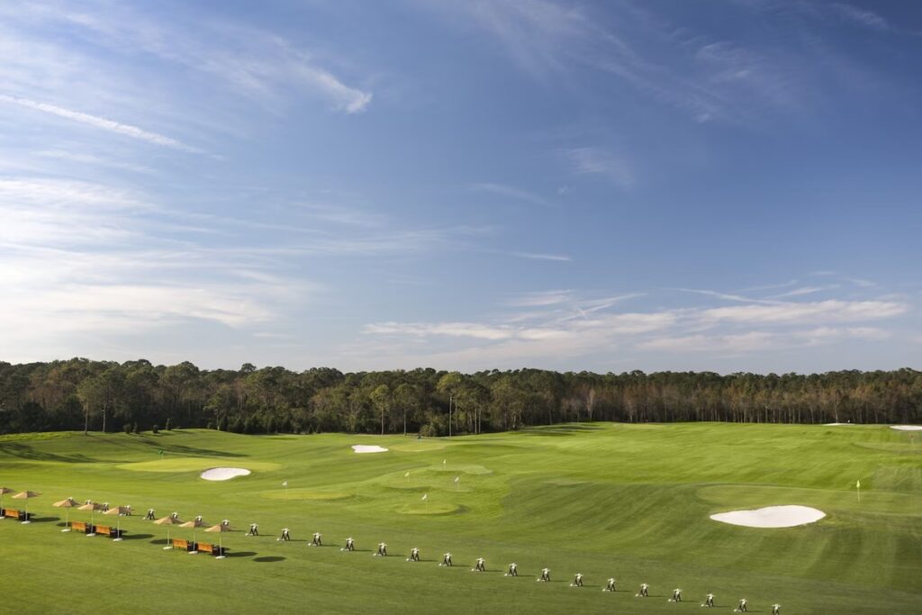 A vast, green golf driving range lined with target umbrellas under a clear blue sky.