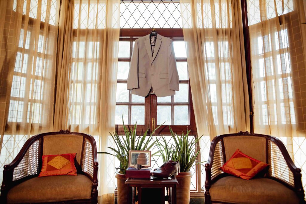 A gray tailored suit jacket hanging on a contoured wooden hanger by a hotel window to naturally release wrinkles after unpacking.