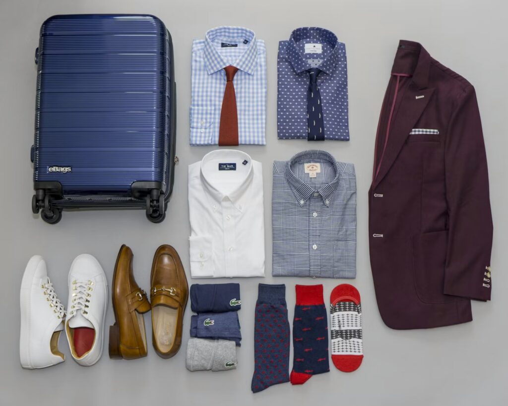 A flatlay of a blue hardshell carry-on luggage next to a burgundy suit, dress shirts, and leather shoes, demonstrating how to pack a suit in a suitcase.